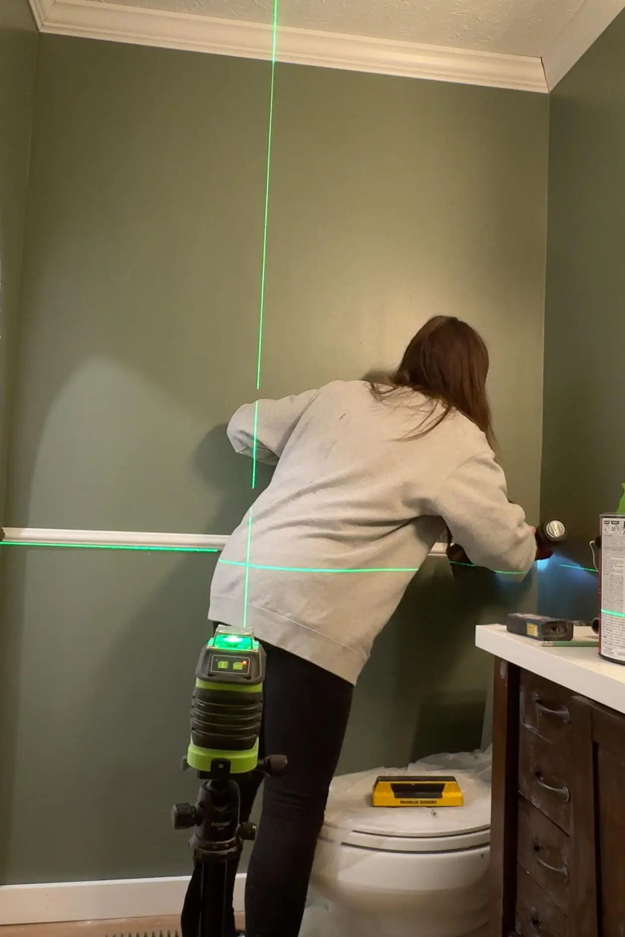 Person aligning chair rail molding on a green bathroom wall with the aid of a laser level, capturing the detailed steps of a DIY picture frame molding project.